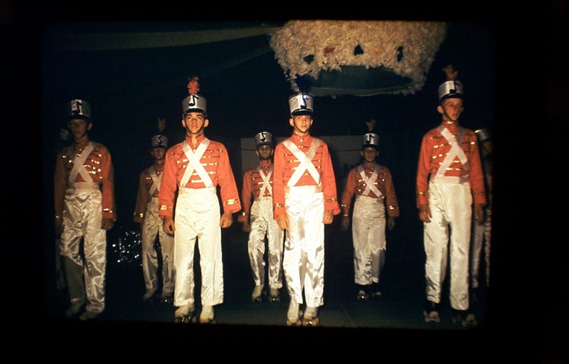 Six men in red coats and white sashes stand in formation at night under a canopy, wearing tall dark helmets.
